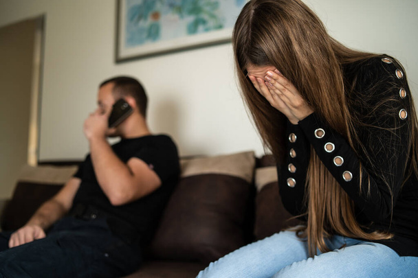 Side view on sad woman crying with hands over her face while unknown man sitting by her on sofa at home or hotel making a phone call talking - human trafficking and prostitution or violence concept - Photo, Image