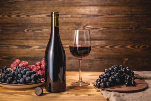 Uma garrafa de vinho tinto seco com um copo e um monte de uvas em uma mesa de madeira. Conceito de viticultura e vinificação - Foto, Imagem