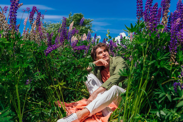 Tall handsome man in a green jacket sitting among the lupine flowers, enjoing the beauty of nature. Man surrounded by purple and pink lupines. - Photo, Image
