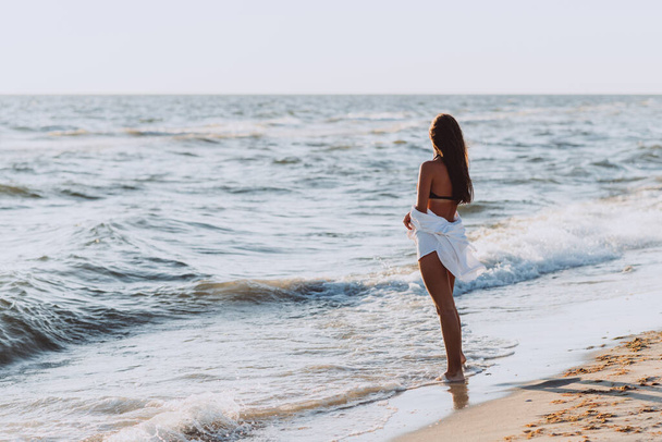 Young beautiful slender woman in a black bikini and white shirt on a tanned body on the beach in the waves, back view. Soft selective focus, art nose. - Photo, Image