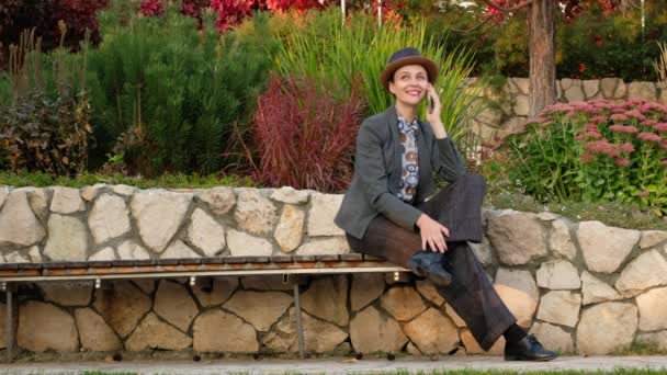 Mujer de negocios con traje y sombrero hablando por teléfono sentada en un banco en el parque. - Imágenes, Vídeo