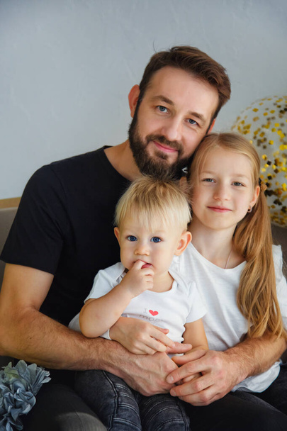 Portrait of a happy father with adorable children sitting on the sofa posing for a family photo at a holiday home. Cuddles with cute little son and daughter while looking at the camera - Photo, Image