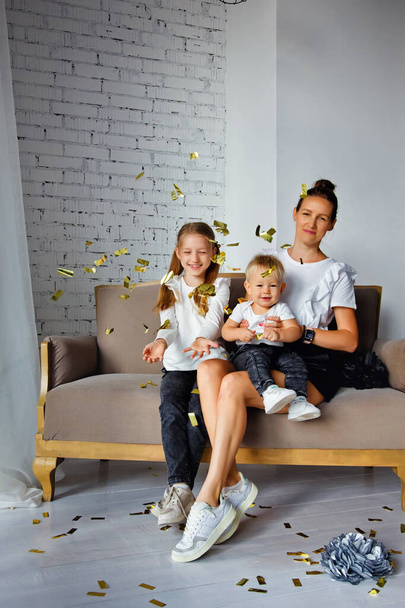 Портрет счастливой матери с очаровательными детьми, сидящими на диване и позирующими для семейного фото в доме отдыха. Обнимается с симпатичными маленькими сыном и дочерью, глядя в камеру - Фото, изображение