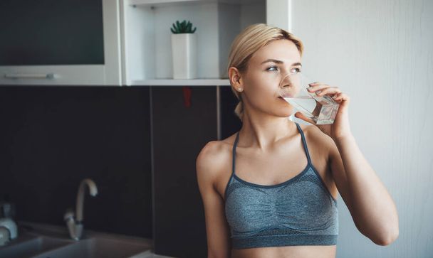 Mooie blanke vrouw met blond haar in sportkleding drinkt een glas water tijdens de digitale fitnesslessen thuis - Foto, afbeelding