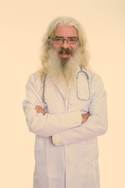 Studio shot of handsome senior man doctor with long white curly beard and hair isolated against white background - Fotó, kép
