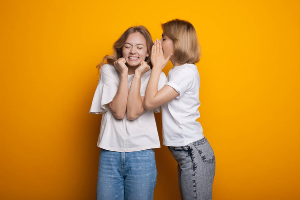 Blonde caucasian woman is whispering something to her friend posing in casual clothes on a yellow studio wall - Photo, Image