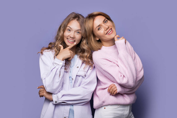 Sarışın kız kardeşler günlük kıyafetleriyle kameraya gülümsüyor menekşe bir stüdyo duvarında çenelerine dokunuyorlar. - Fotoğraf, Görsel