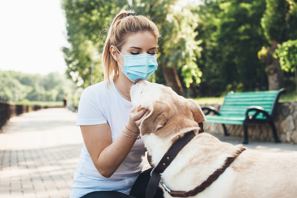 Lovely blonde girl and her labrador are embracing in a park while wearing a medical mask - Photo, image