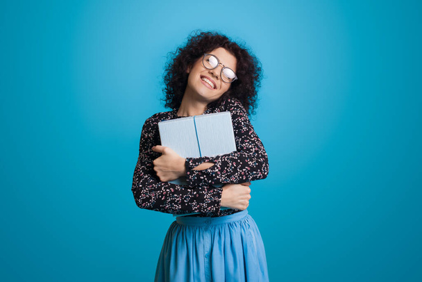 Dulce mujer caucásica con el pelo rizado y gafas está abrazando una caja de regalo sonriendo en una pared de estudio azul en un vestido - Foto, Imagen