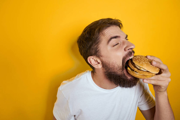 Man with hamburger in the hands of fast food διαίτης λευκό t-shirt close-up κίτρινο φόντο - Φωτογραφία, εικόνα