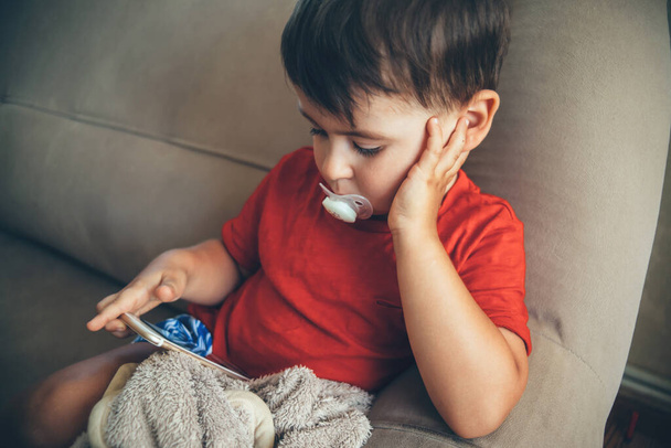 Piccolo ragazzo caucasico seduto sul letto e che guarda lo schermo del telefono mentre tiene un alimentatore in bocca - Foto, immagini