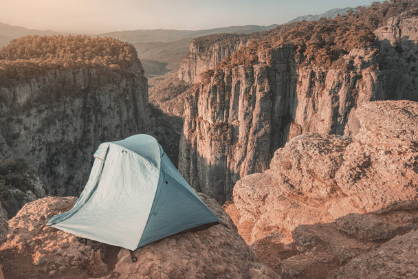 Een eenzame blauwe tent ligt bovenop een klif boven een diepe kloof met een prachtig uitzicht op de bergen. Concept van extreme rotsklimmen en kamperen op gevaarlijke plaatsen - Foto, afbeelding