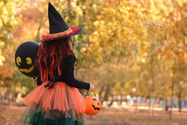 Little girl in witch costume with a balloon and a pumpkin bucket playing in autumn park. Child having fun at Halloween trick or treat. Kids trick or treating. - Photo, Image