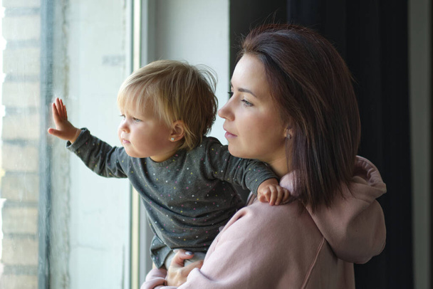 Tourné de jeune mère heureuse avec une petite fille regardant par la fenêtre à la maison. - Photo, image