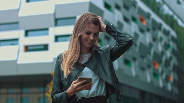 Good looking girl with Long Brown Hair Looking at Her phone Typing and Smiling. Enormous industrial Building at the Background. Green Bushes and Trees. Smart clothes. Natural makeup. - Photo, Image