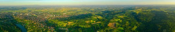 Above view, wide skyline panorama, over green hilly landscape, several cultivated, arable plots, among forest trees and city in the distance. - Photo, Image