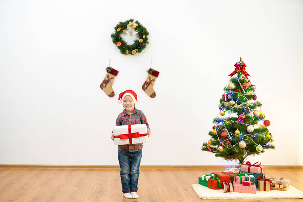De gelukkige jongen staat bij de kerstboom en houdt een geschenkdoos vast - Foto, afbeelding