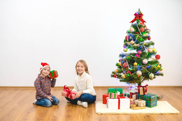 Το χαρούμενο αγόρι και ένα κορίτσι κάθονται με κουτιά δώρων κοντά στο χριστουγεννιάτικο δέντρο. - Φωτογραφία, εικόνα