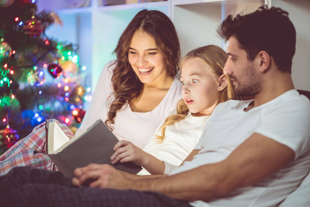 Счастливая семья лежала на кровати и читала книгу возле рождественской елки - Фото, изображение
