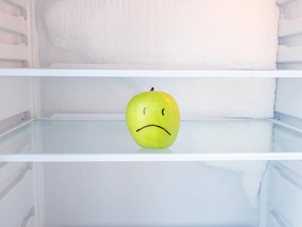 Ένα πράσινο λυπημένο μήλο σε ανοιχτό άδειο ψυγείο με παγωμένο πάγο στον τοίχο. δίαιτα απώλειας βάρους και την έννοια της πείνας. - Φωτογραφία, εικόνα