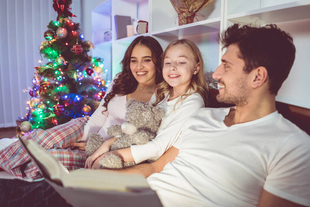 幸せな家族はベッドの上に横たわってクリスマスツリーの近くの物語を読む - 写真・画像