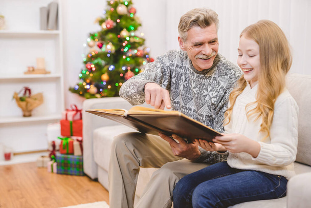 A menina com um avô olha para o álbum de fotos perto da árvore de Natal - Foto, Imagem