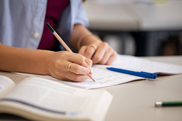 Close up of college student hand on desk in classroom writing notes. Closeup of university girl hands taking notes from textbook during lecture. Detail of young woman using pencil on notebook in classroom during lesson at high school. - 写真・画像