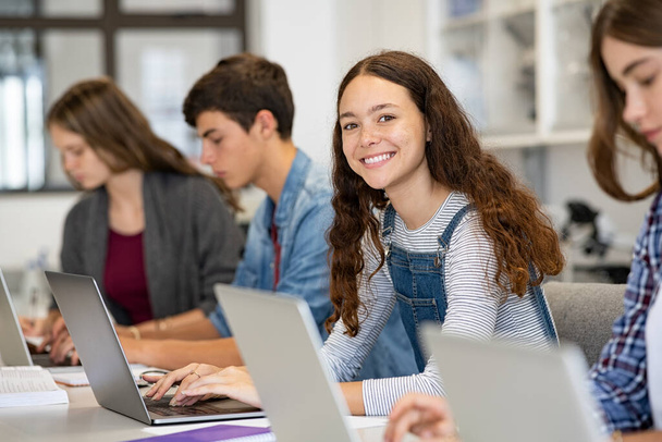Happy young woman working on laptop and looking at camera in classroom. Portrait of smiling university student in library use computer for a research. Satisfied college student looking at camera while sitting in a row with classmates studying togethe - 写真・画像
