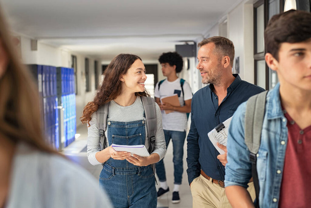 Happy high school student talking to mature man professor in hallway. Senior man teacher in conversation to school girl holding book in campus. Helpful lecturer explaining concept to college student outside classroom. - Фото, зображення