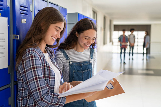 Best friends sharing notes standing near locker in college campus. Happy smiling university students laughing and holding notebook discussing notes. Two beautiful girls laughing and talking after class at high school hallway with copy space. - Foto, Bild