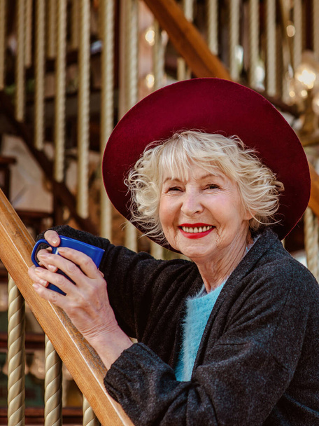 senior stylish woman in dark grey coat, hat and with grey hair standing by the carousel smiling, drinking coffee (tea) and enjoying life. Travel, fun, pension, happiness, seasonal concept - Foto, Imagem