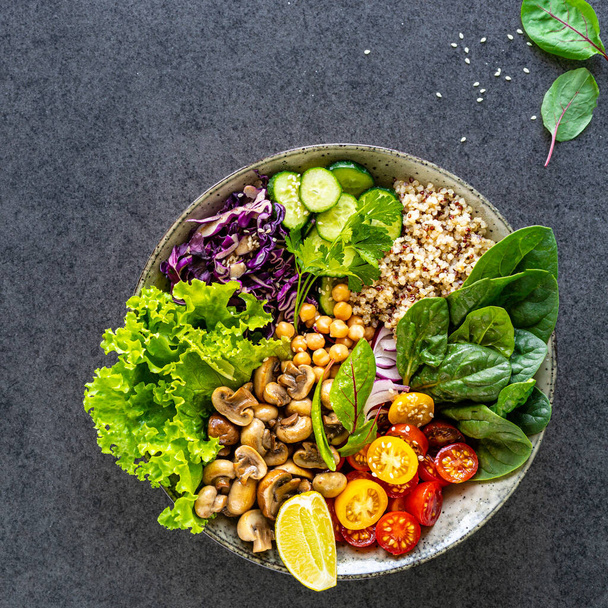 Quinoa, mushrooms, lettuce, red cabbage, spinach, cucumbers, tomatoes, a bowl of Buddha on a dark background, top view. Delicious balanced nutrition concept - Photo, Image