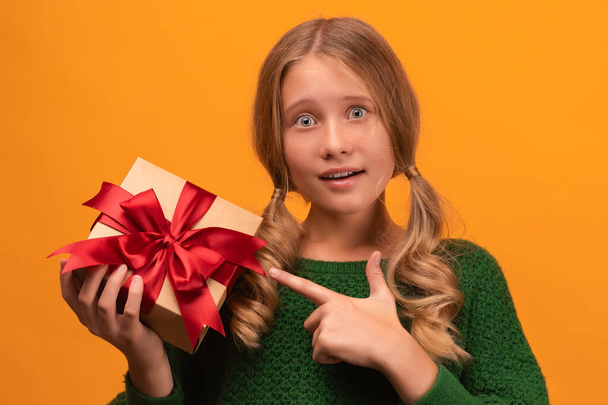 Image of charming blonde girl 12-14 years old in warm green sweater holding present box with red bow. Studio shot, yellow background, isolated. New Year Women's Day birthday holiday concept - Valokuva, kuva