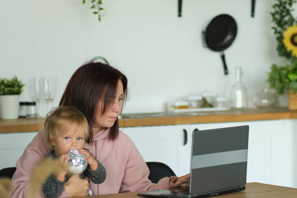 mom working remotely on laptop while taking care of her baby. Young mother on maternity leave trying to freelance by the desk with toddler child. Close up, copy space, background. - Fotografie, Obrázek