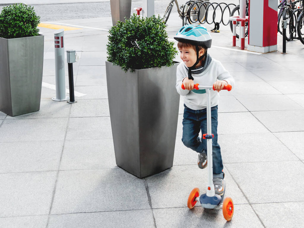Toddler in helmet is riding scooter on parking lot with parked bicycles. Urban vehicle for active children. Leisure activity for boys and girls. - Foto, imagen