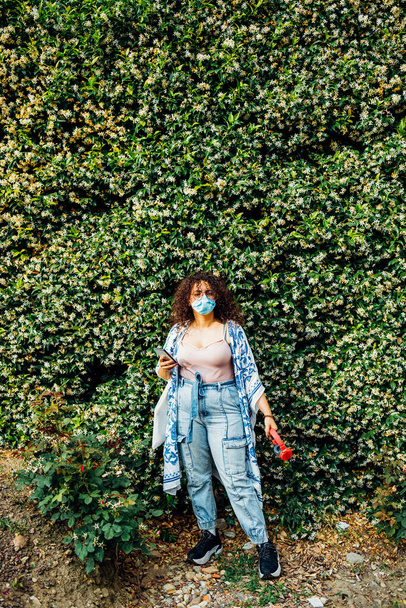 Junge multiethnische Frau posiert im Freien mit Schutzmaske und Handy - Gemischte Rasse junge Frau trägt Gesichtsmaske mit Smartphone in der Stadt - Schutz, Umweltverschmutzung, Technologiekonzept - Foto, Bild