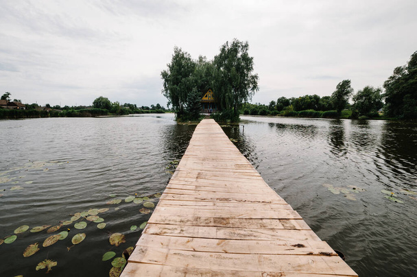 Bridge, masonry across through the river. Close up. A young couple standing near a wooden old house on an island in the middle of a lake. House on the background of trees. Nature, landscape. - Фото, изображение