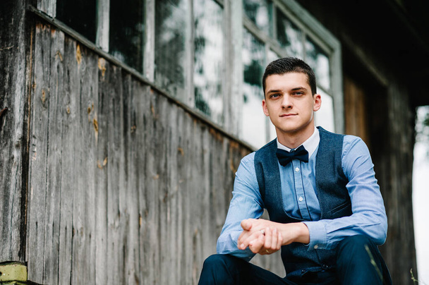 Portrait young beautiful man sitting next to the wooden wall of an old house. looking at camera. upper half length. businessman. Close up. - 写真・画像