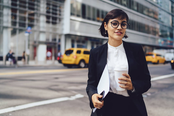 Selbstbewusste Managerin in schwarzem Anzug und Brille, die auf der Straße steht und Kaffee zum Mitnehmen trinkt, während sie in die Kamera blickt und Smartphone und Dokumente in der Hand hält in New York City - Foto, Bild