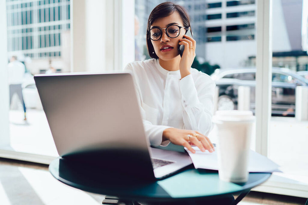 Serious ethnic female in formal clothes and eyeglasses sitting in modern office while working on laptop and talking on smartphone - Фото, зображення