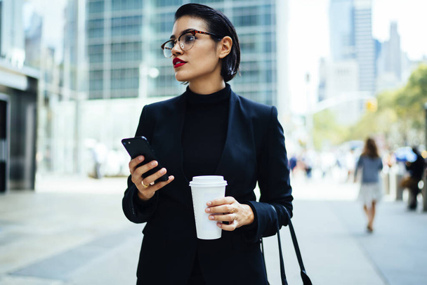 Serious young businesswoman in elegant outfit and glasses holding smartphone and cup of hot drink standing on steet in downtown of New York City and looking away - Fotografie, Obrázek