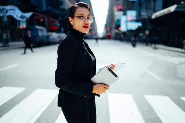 Female manager with short hair and red lips in total black outfit holding documents and tablet in middle of crosswalk - Foto, Imagem