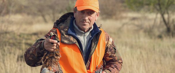 Un uomo con una pistola in mano e un giubbotto arancione a caccia di fagiani in una zona boscosa con tempo nuvoloso. Un cacciatore con un fagiano in mano. - Foto, immagini