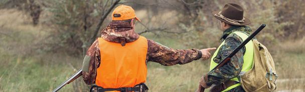 A mans with a gun in his hands and an orange vest on a pheasant hunt in a wooded area in cloudy weather. Hunters with dogs in search of game. - Photo, Image