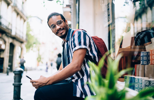 Half length portrait of smiling dark skinned male millennial using smartphone communicating and browsing on free time, smiling african american looking at camera satisfied with 4G connection on street - Foto, immagini