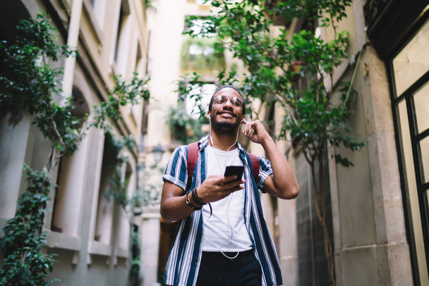 Low angle of joyful ethnic guy in casual summer outfit and eyeglasses using headset for talking on smartphone while strolling in historical part of city - Photo, Image