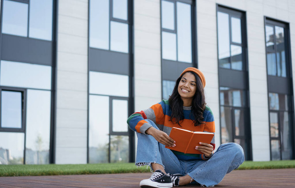 Portrait of young beautiful African American woman reading book outdoors. Happy university student studying, learning language, exam preparation, education concept - Photo, Image