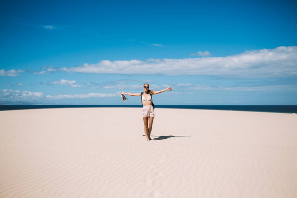 Blond female traveler in swimsuit enjoying time on sandy beach against blue sea of island while holding hands apart and smiling  - Fotografie, Obrázek