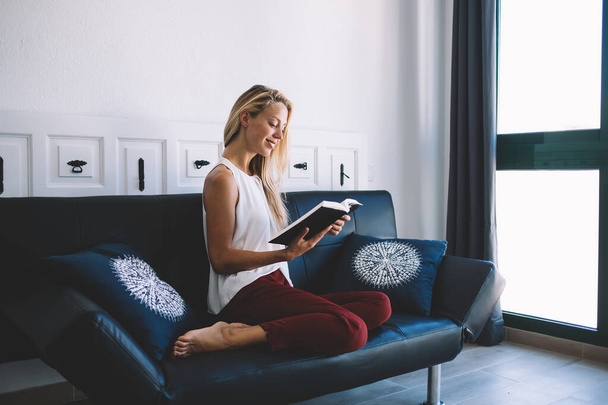 Beautiful woman dressed in casual look resting at comfortable couch and reading interesting bestseller about youth lifestyle, young hipster girl relaxing during literature hobby in home interior - Fotografie, Obrázek