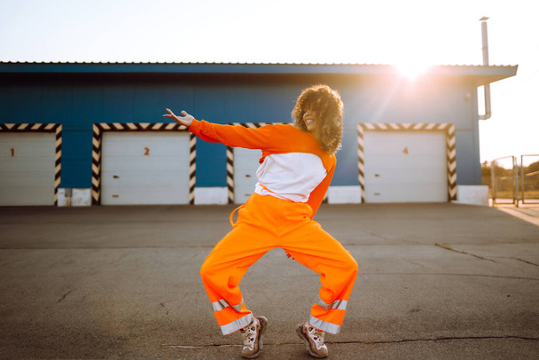 Young sporty woman dancer dancing in the street at sunset. African American woman in an orange suit showing some moves. Sport, dancing and urban culture concept. - Fotografie, Obrázek